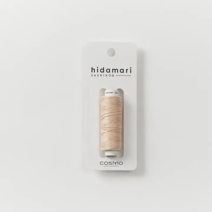 A spool of light beige sewing thread mounted on a white card packaging, featuring the brand names 'hidamari' and 'cosmio' on a clean white background.